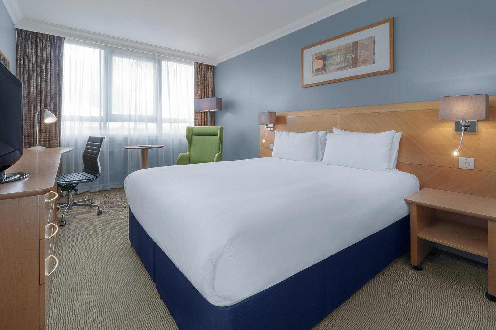 A guest room at Holiday Inn London – Kensington Forum: a double bed, a desk and a green armchair by a wide window